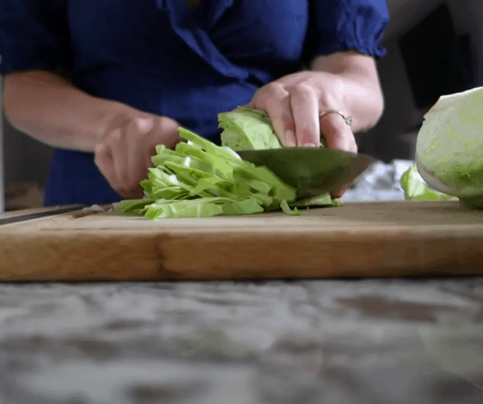 bright green cabbage cut into shreds on wooden cutting board with woman cutting using a chefs knife in a blue dress