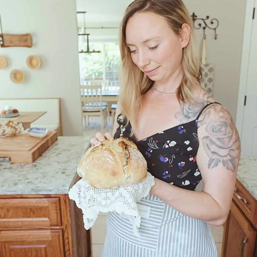 Kyrie from healthfully rooted home holding loaf of roasted garlic sourdough bread on top of a lace doily wearing a floral dress with aprons in the background.
