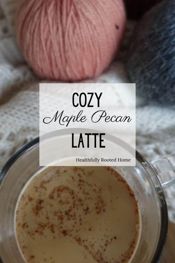 Fall coffee drink maple pecan latte with coffee in a glass coffee cup on top of a white cozy sweater with pink and blue sweater pumpkins in back