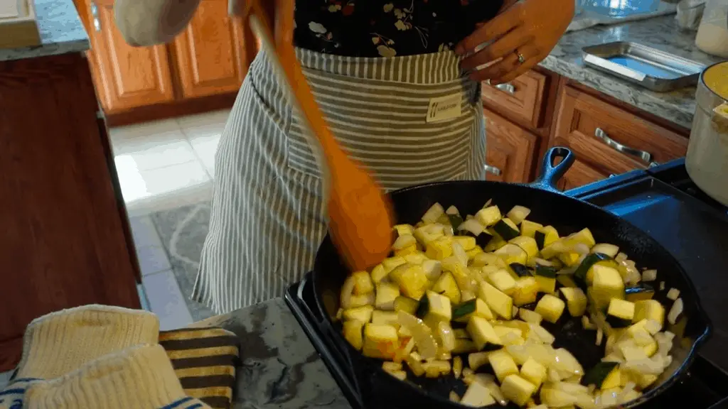 sauteeing zucchini and onions in a cast iron skillet with a wooden spoon wearing striped apron