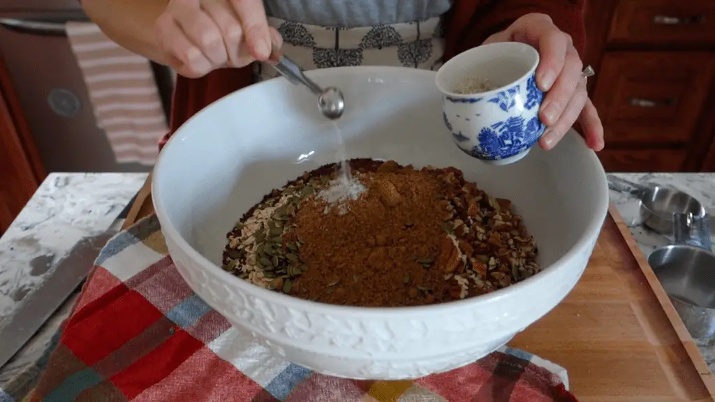 mixing the dry ingredients for pumpkin spice granola bark in a large white bowl on top of a boos block wooden butcher block sing stainless steel measuring spoons with a red checkered tea towel