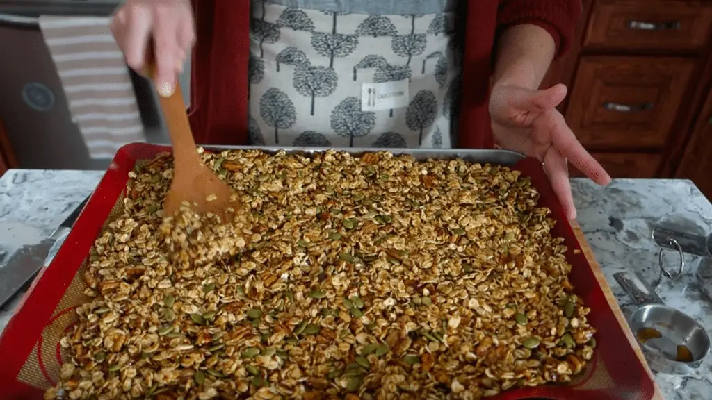 spreading pumpkin spice granola bark on a silicone mat lined baking sheet using a wooden spoon with stainless steel measuring cups to the side