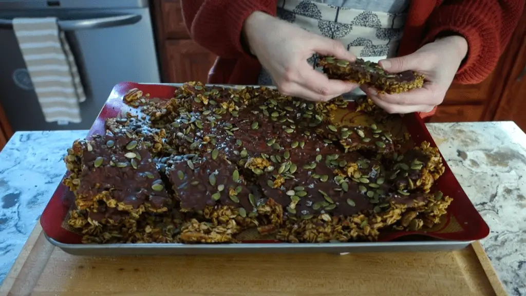 breaking apart the pumpkin spice granola bark on a silicone mat lined baking sheet wearing a chunky red sweater and an apron with trees on it