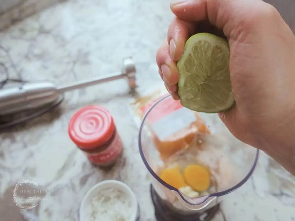 squeezing the juice out of a lime into an immersion blender with harissa paste and chili powder in the background