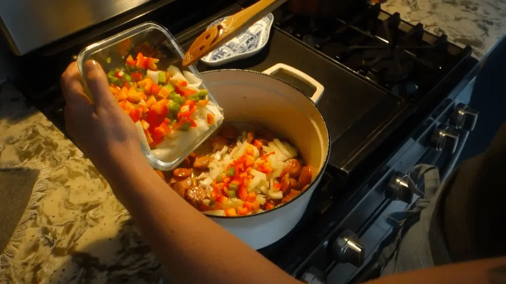 adding vegetables to the sausage in a white dutch oven