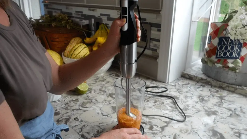 using an immersion blender to blend ingredients inside an immersion cup with a fruit bowl in background