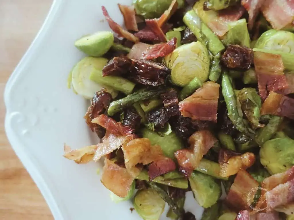 roasted asparagus and brussels sprouts with dates and bacon on a white serving tray and boos block cutting board in background