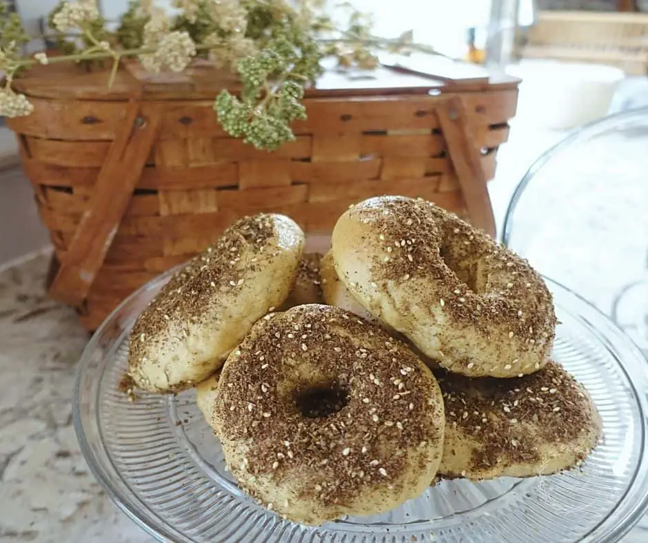 homemade sourdough bagels with zaatar seasoning on a glass clear cake stand with a basket in the background on a granite countertop