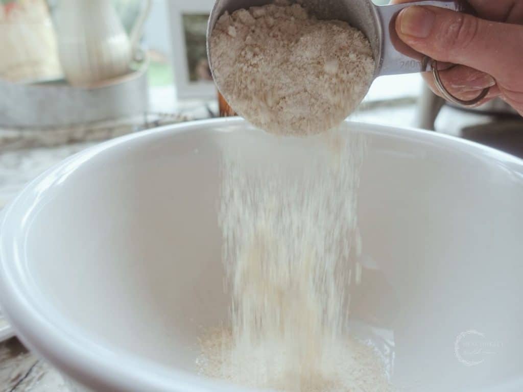 pouring freshly milled einkorn flour into a ceramic bowl using a stainless steel measuring cup