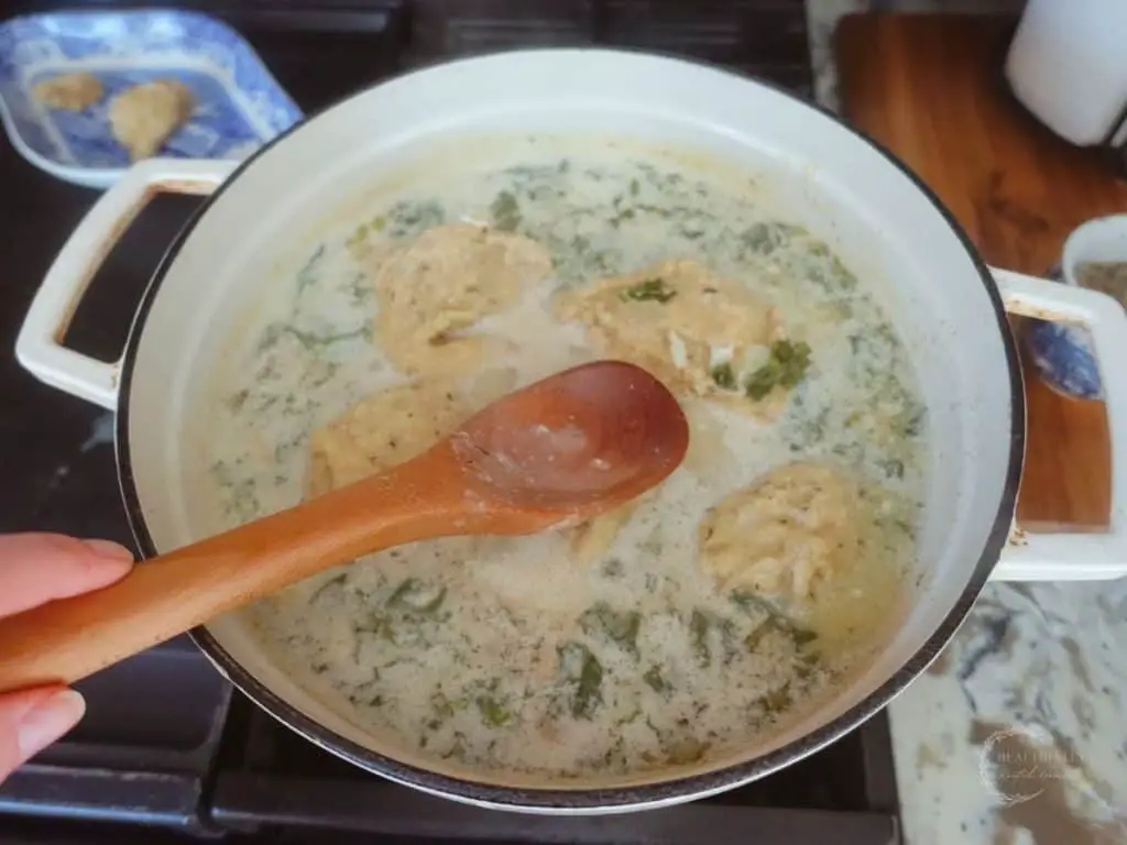 using a wooden spoon to push down drop sourdough dumplings into a dutch oven full of chicken kale soup