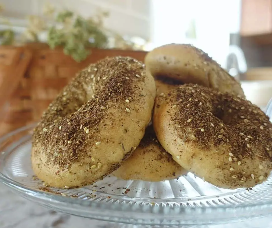 homemade zaatar bagels on a glass cake stand with a brown basket in the background with green and white stems