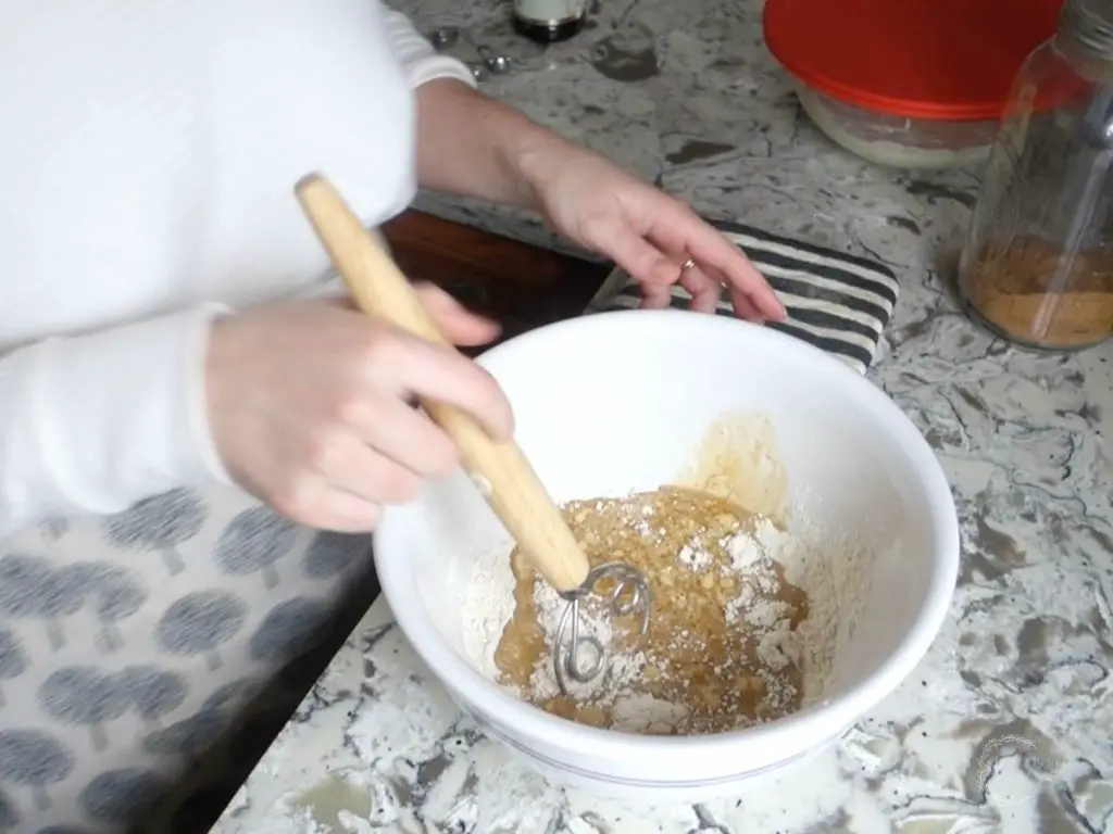 combining wet and dry ingredients with a danish dough whisk in a large ceramic bowl wearing a white sweater and apron
