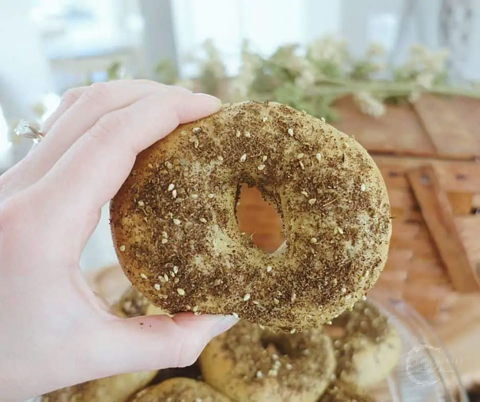 sourdough bagel with zaatar seasoning on top held up between two fingers with a basketweave basket in the background with green and cream stems on top and a batch of sourdough bagels on a cake stand
