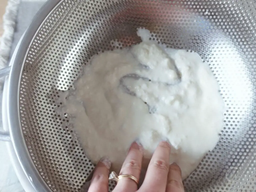 straining raw kefir in a metal strainer using fingers wearing a gold wedding ring