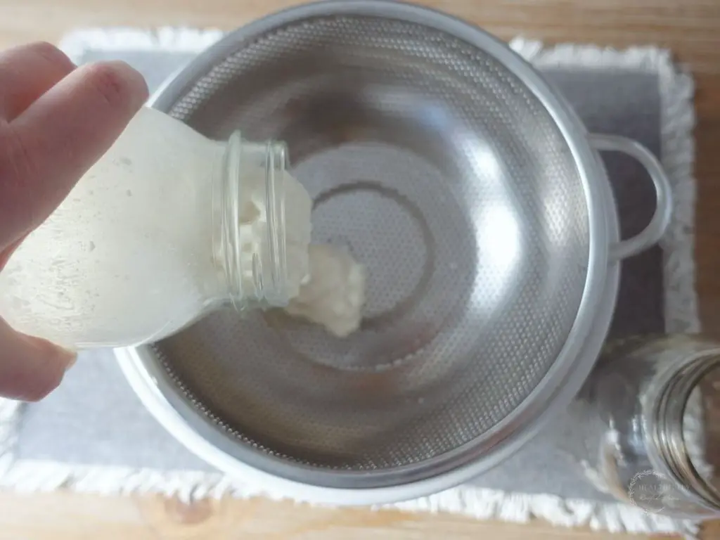 pouring raw kefir into a strainer with a mason jar to the side on top of a grey placemat