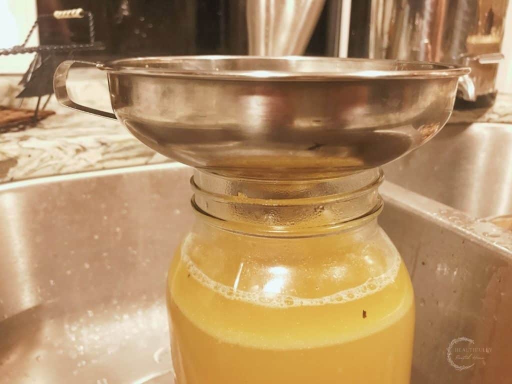 stainless steel funnel over a half gallon mason jar inside a stainless sink with chicken collagen soup inside