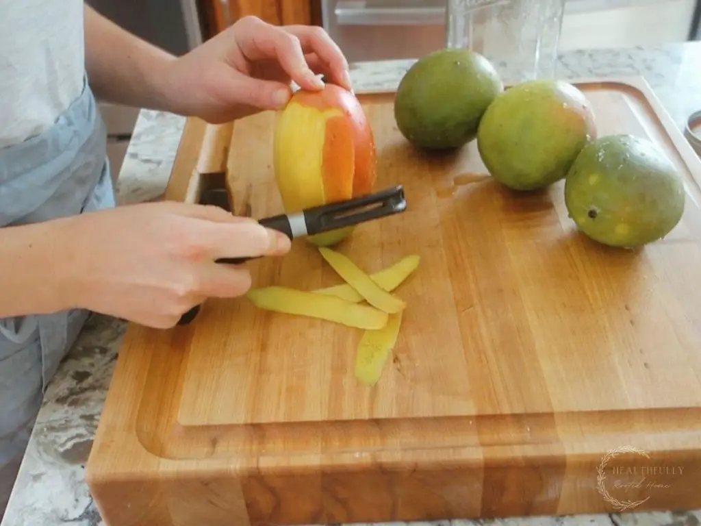peeling a mango on a wooden boos block cutting board with 3 other mangos in the background