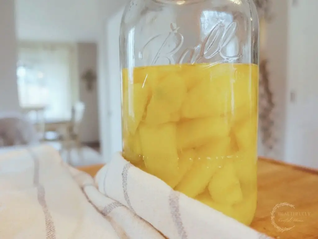 fermented mangos in a large mason jar next to a blue and white striped tea towel with a cross in the background