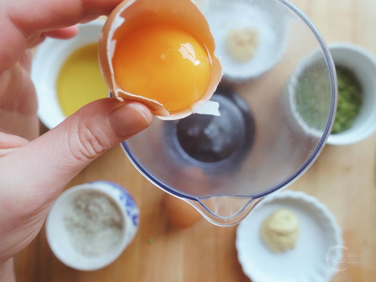 adding an egg yolk to the cup of an immersion blender with various ingredients in white cups surrounding