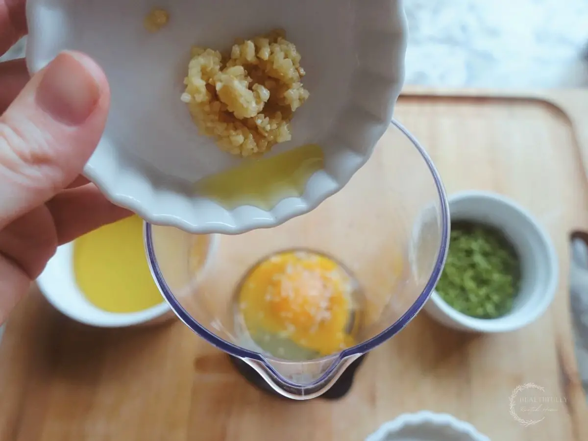 adding garlic into the cup of an immersion blender with eggs inside