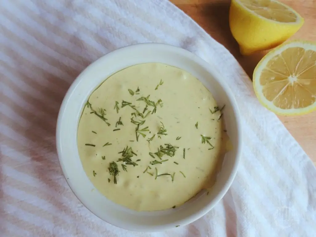 dill aioli in a white container with lemons on the side on top of a white and cream colored ticking stripe tea towel