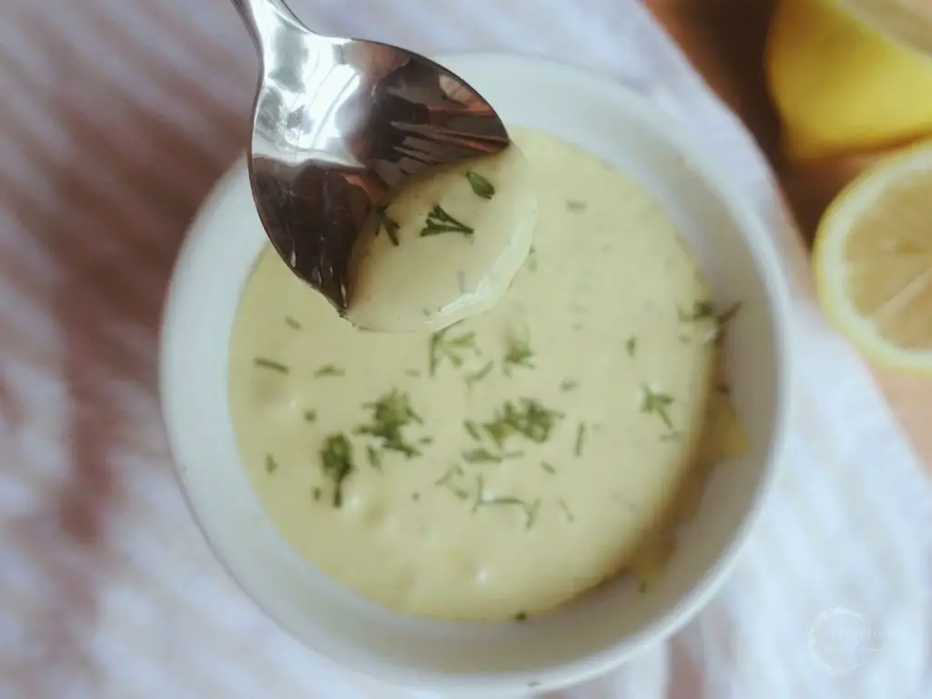 dill aioli on a silver spoon with fresh dill as garnish on top of a white and brown ticking stripe tea towel