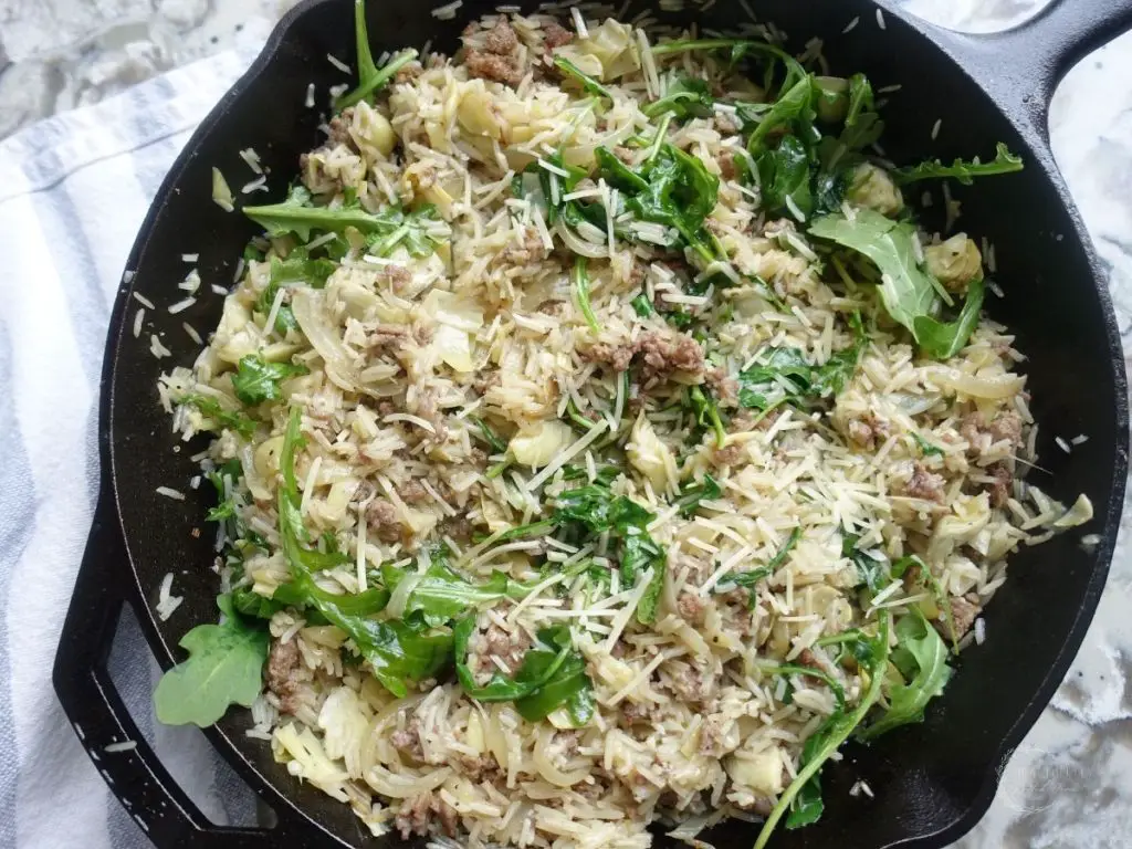 risotto basmati with Italian sausage and artichokes and arugula inside of a cast iron skillet with a ticking stripe tea towel beside it