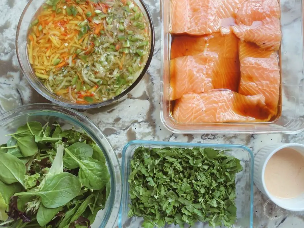 ingredients for salmon banh mi salad in clear glass containers on marble countertop with raw salmon pickled veggies spring greens cilantro and ancho aioli