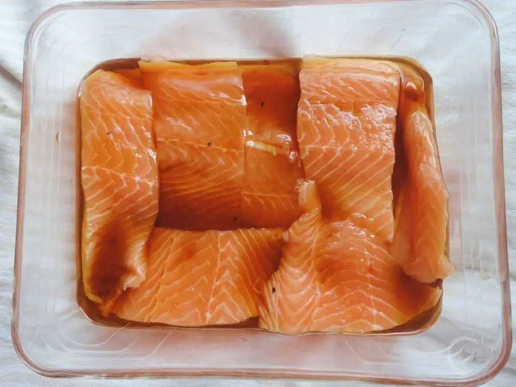 salmon marinating in sesame oil in a glass baking dish overtop a white and cream ticking stripe tea towel