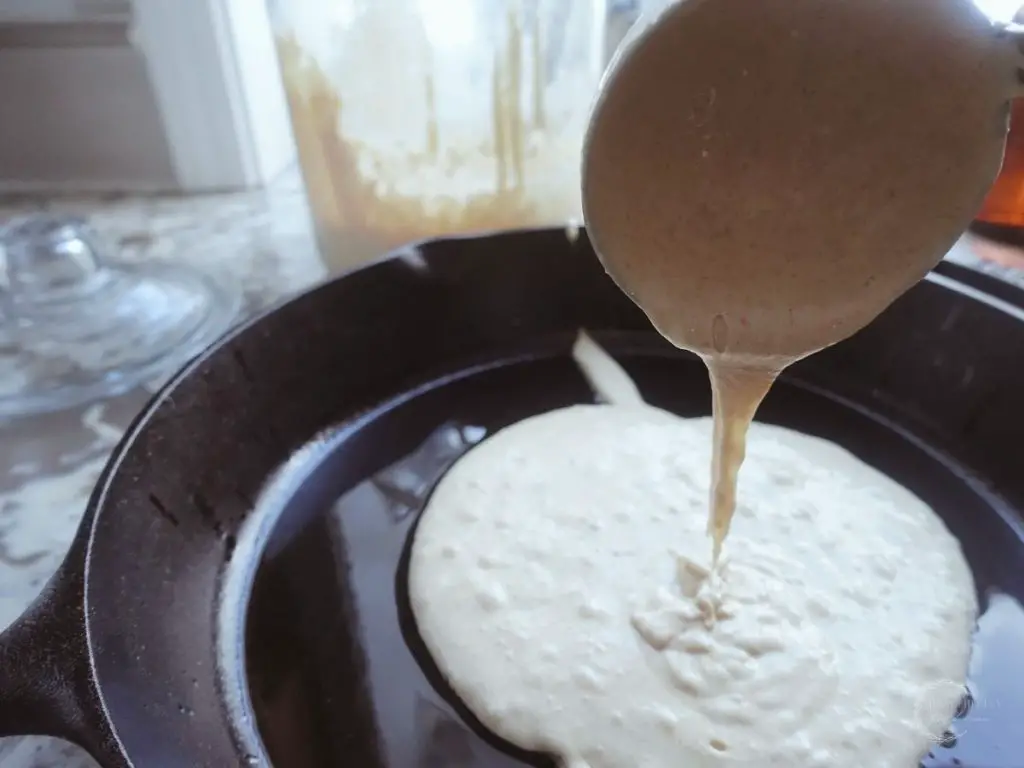 using a ladle to pour sourdough starter on a cast iron skillet with a jar of sourdough starter in the background