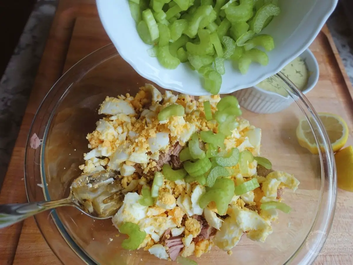 pouring chopped celery into a bowl with eggs and tuna salad