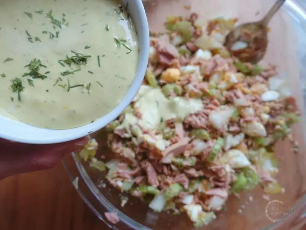 pouring dill aioli over the mixture of tuna salad in a glass bowl