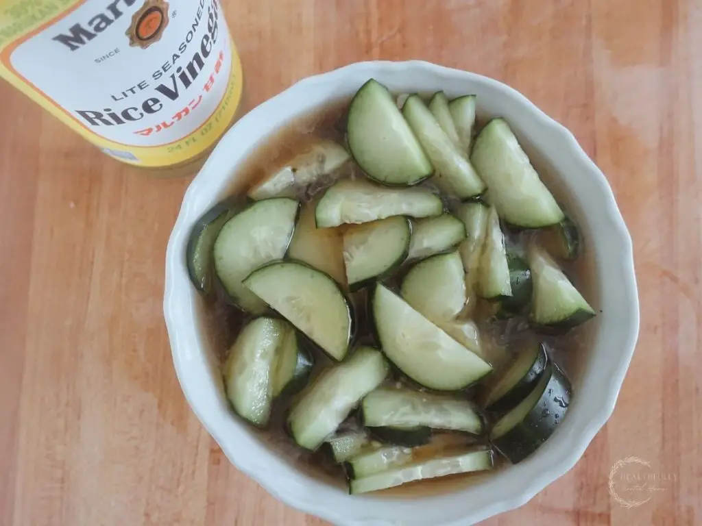 quickly pickled cucumbers in a white bowl with rice vinegar on the side sitting on a wooden cutting board