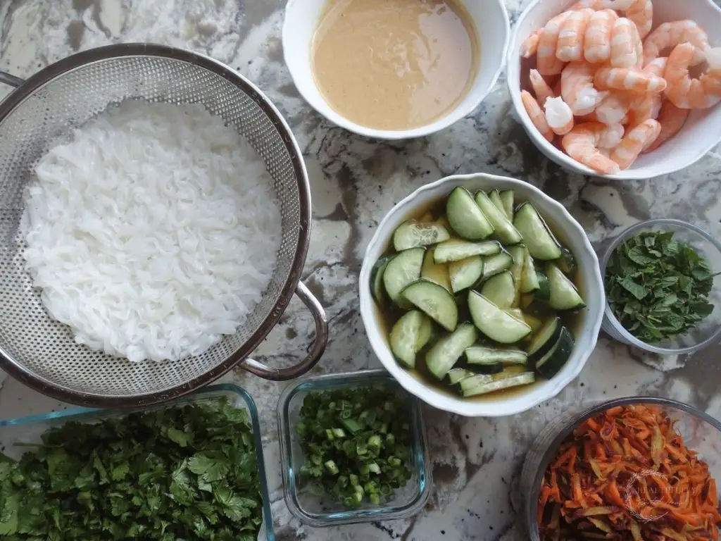 rice noodles in a colander, peanut sauce in a white bowl, pickled cucumbers in a white bowl, shrimp in a white bowl, carrots, mint and cilantro in glass bowls