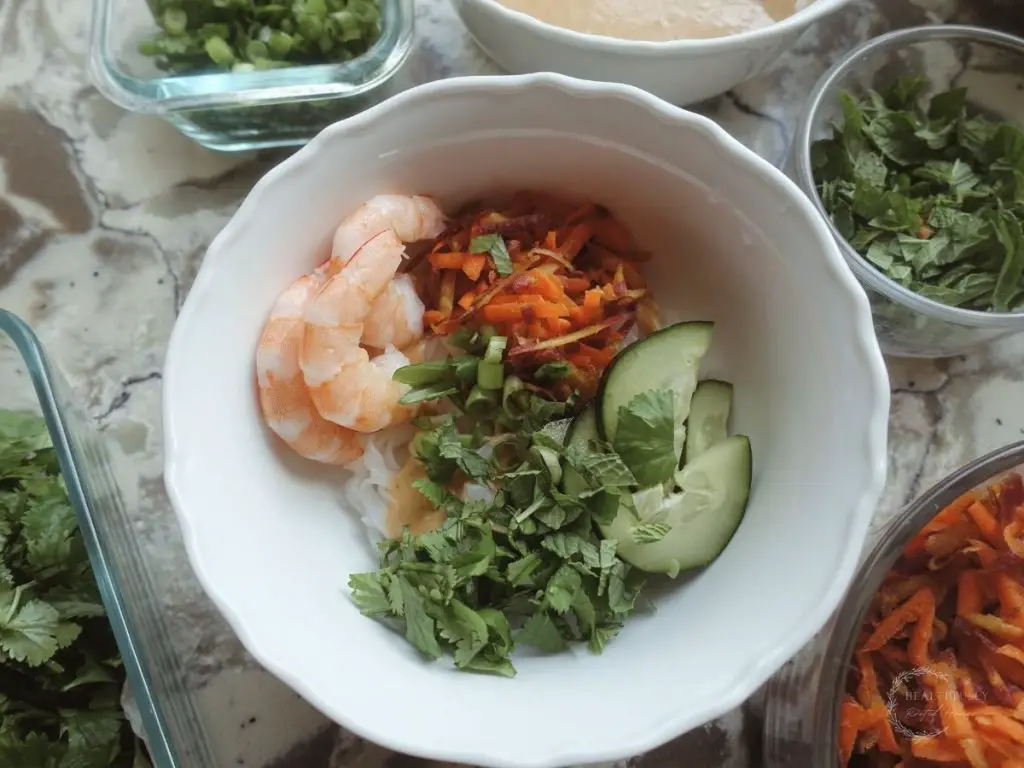 spring roll in a bowl with cucumbers shrimp cilantro carrots and mint all in a white bowl with various ingredients surrounding it