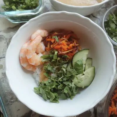 spring roll in a bowl with cucumbers shrimp cilantro carrots and mint all in a white bowl with various ingredients surrounding it