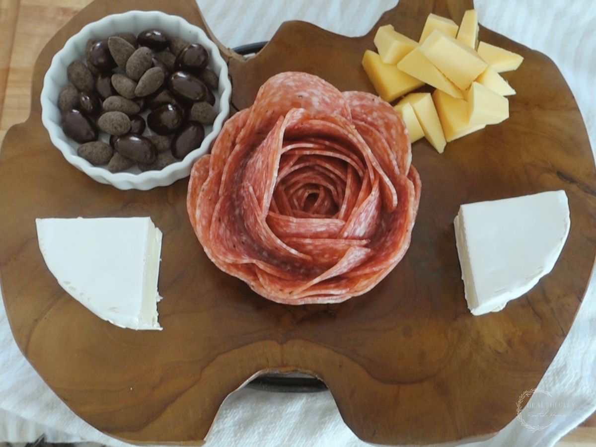 salami rose on a smalll charcuterie board with brie cheese smoked gouda and chocolate covered almonds