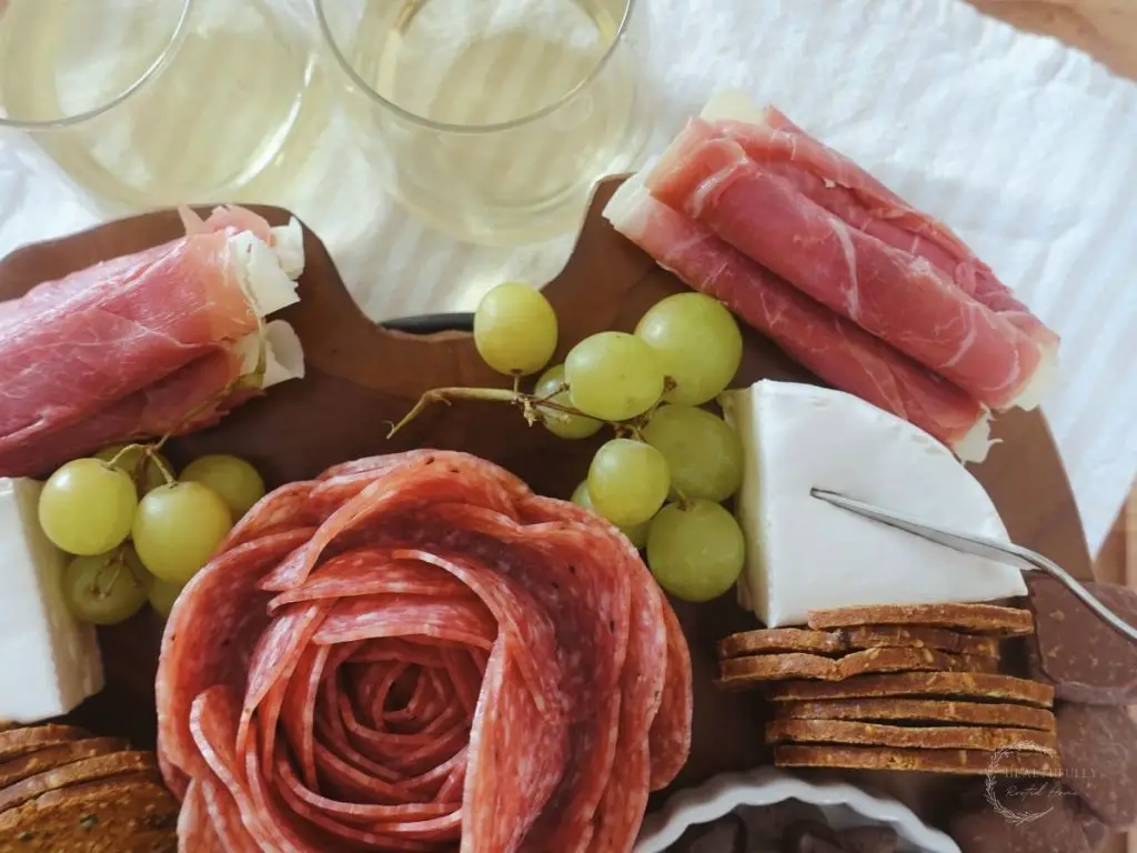 small charcuterie board on a wooden platter with a striped tea towel behind it with two white wine glasses beside it and a salami rose in the center