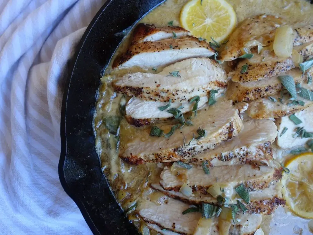 creamy lemon sage sauce for chicken in a cast iron skillet with chicken breasts inside the cream sauce with meyer lemons floating on top