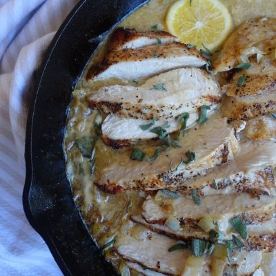 creamy lemon sage sauce for chicken in a cast iron skillet with chicken breasts inside the cream sauce with meyer lemons floating on top