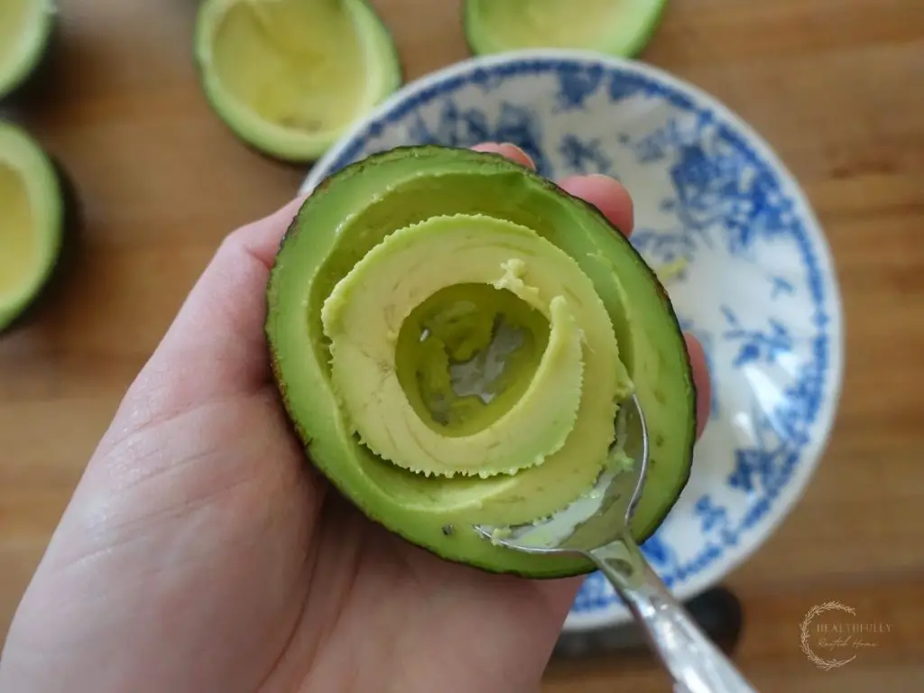 using a spoon to remove some of the avocado fruit from the center of the avocado with a blue and white bowl and avocado halves in the background