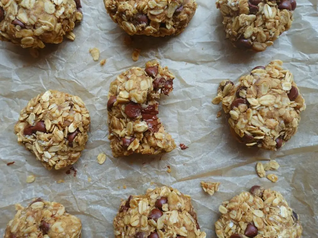 a line of healthy oatmeal cookies on a baking sheet lined with crinkled parchment paper with one of the cookies being bitten