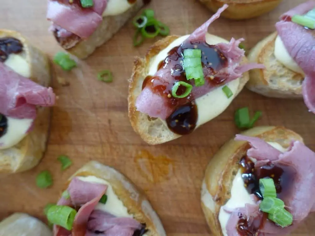 roast beef crostini with horseradish aioli and topped with green onions on top of a wooden cutting board