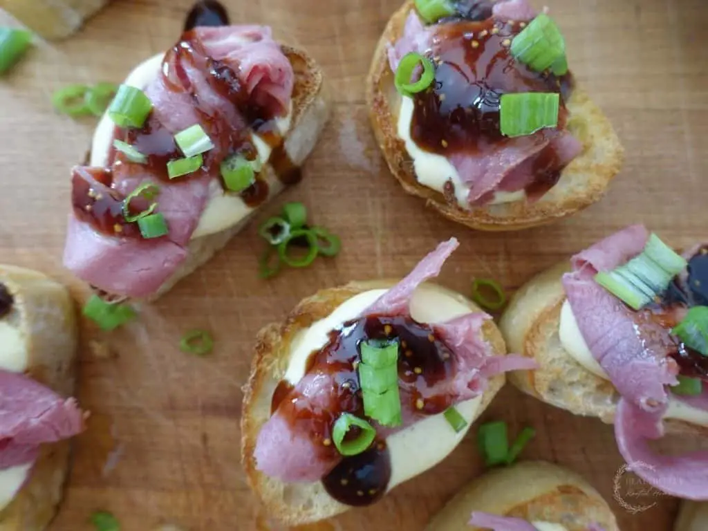 beef crostini topped with balsamic fig glaze and green onions using roast beef on top of a wooden cutting board