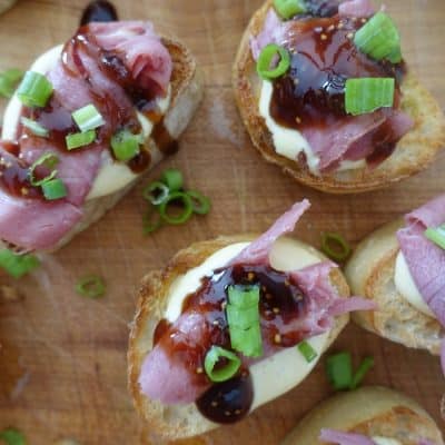 beef crostini topped with balsamic fig glaze and green onions using roast beef on top of a wooden cutting board