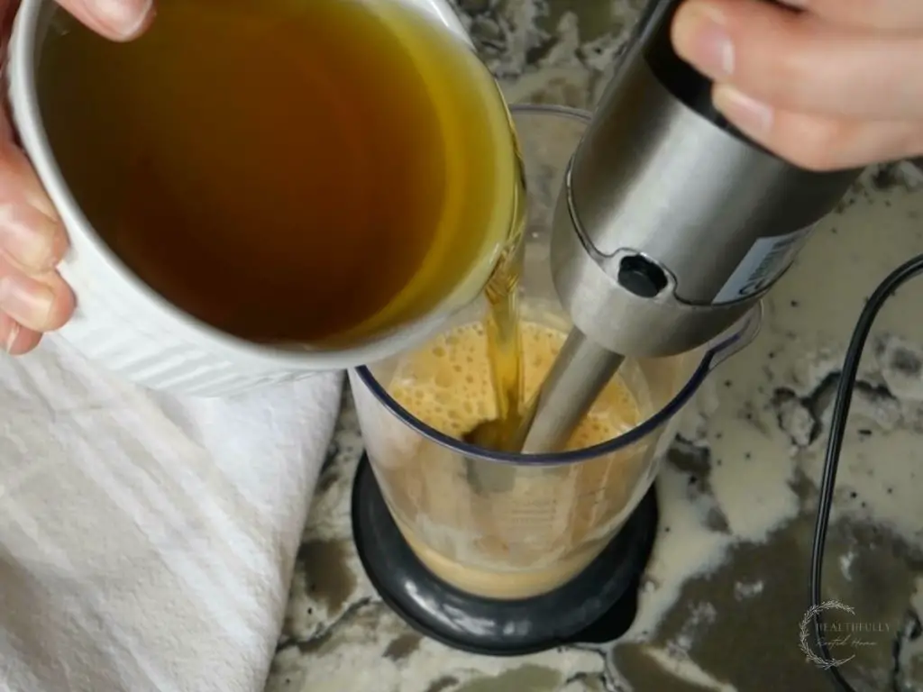 slowly pouring avocado oil into the cup of an immersion blender while also blending