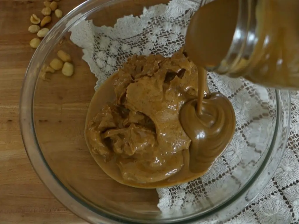 pouring homemade sweetened condensed milk overtop peanut butter in a glass bowl with peanuts and a doily in the background