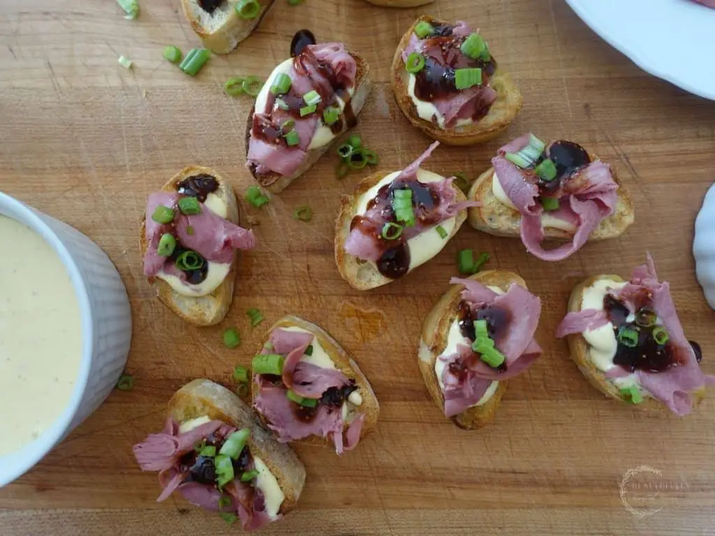 slices of beef crostini on a wooden cutting board with a bowl of horseradish aioli next to them
