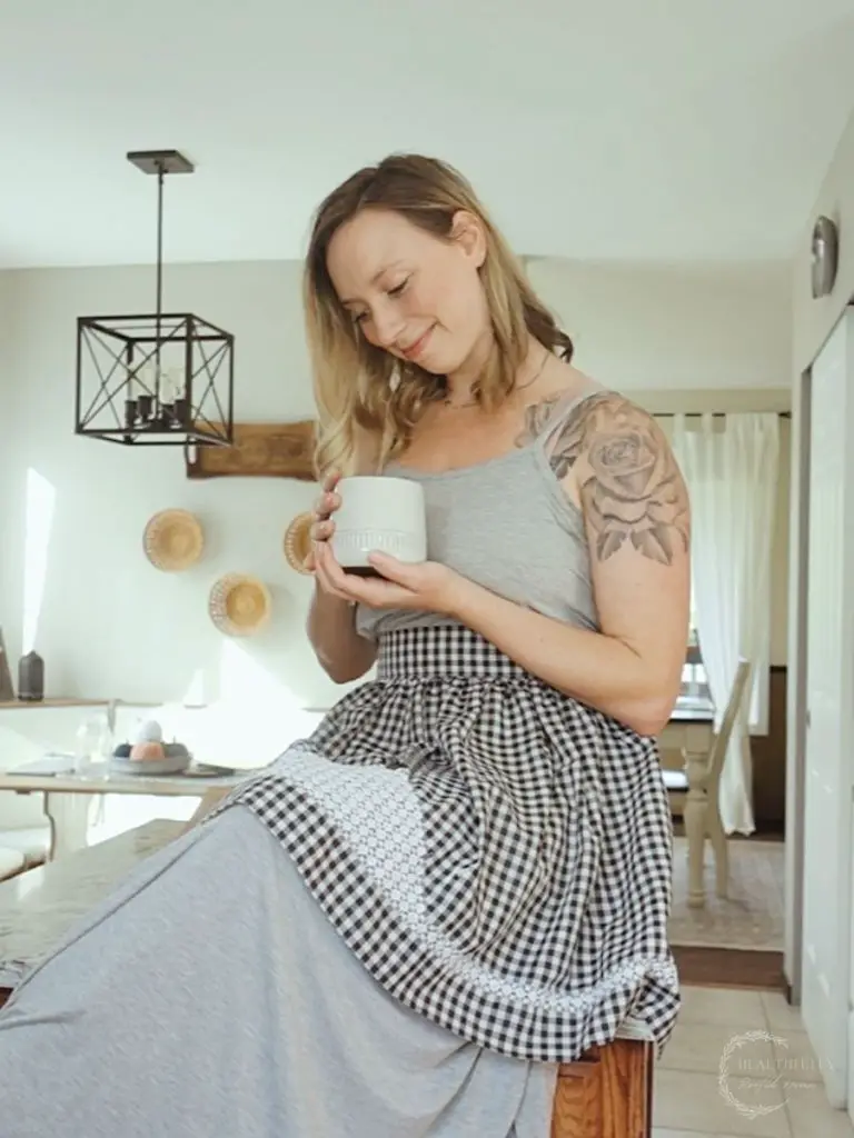 woman with a tattoo wearing a waist apron in a long dress holding a cup of coffee sitting on a kitchen island