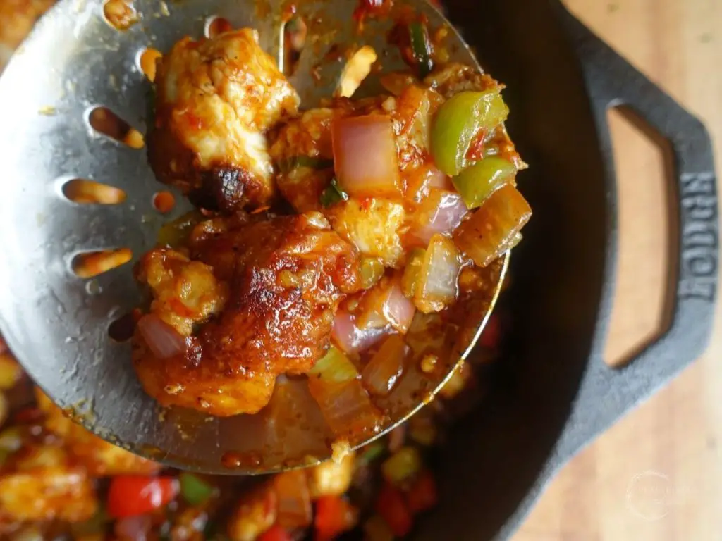 using a fryer spoon to scoop out manchurian chicken out of a cast iron skillet